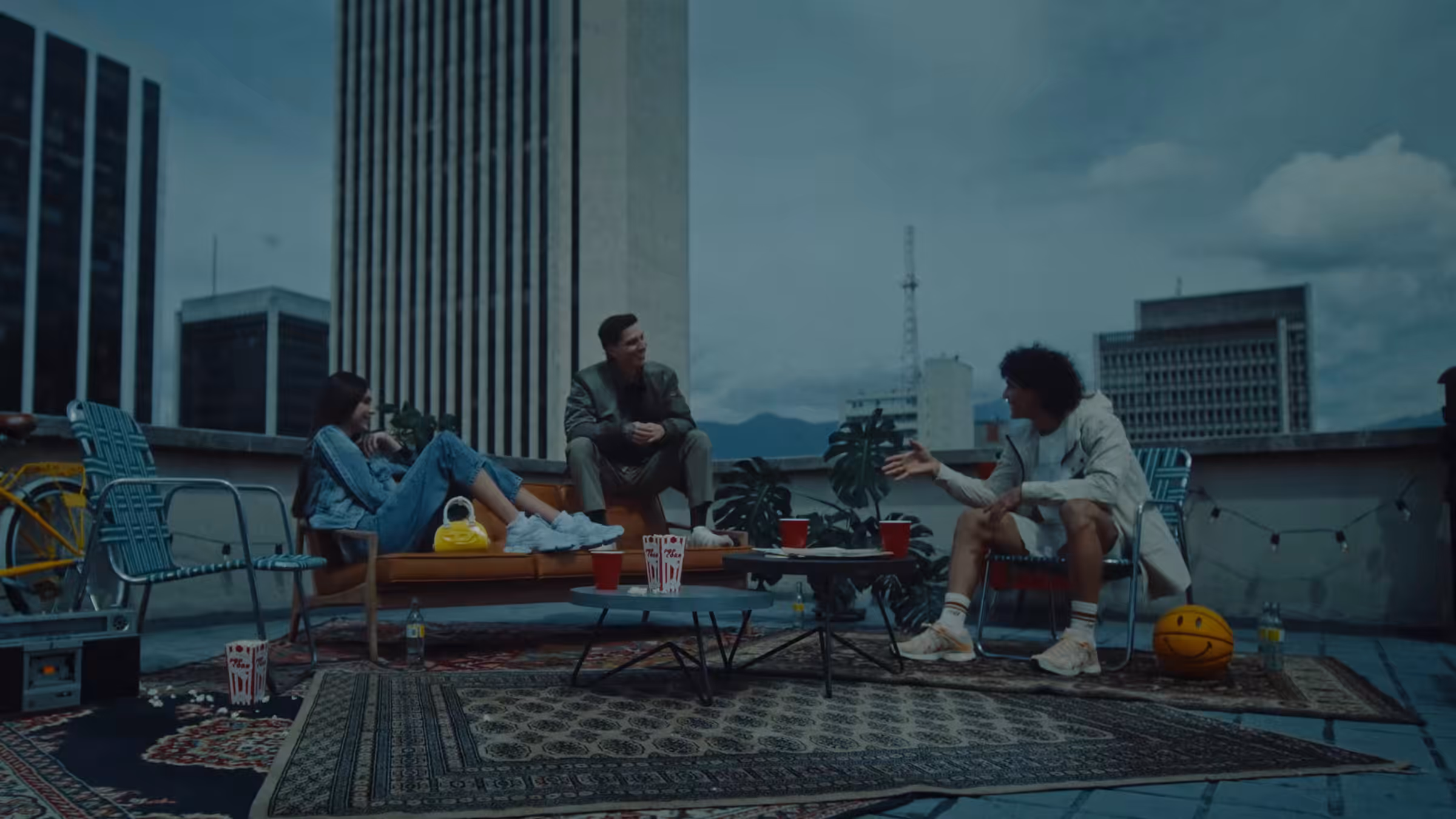 Film still from Pilatos Gravity, three friends on a Medellin rooftop with rugs, folding chairs and the mountains behind them at dusk